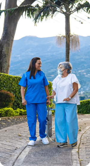 A caregiver walks outdoors with an elderly woman, holding hands.
